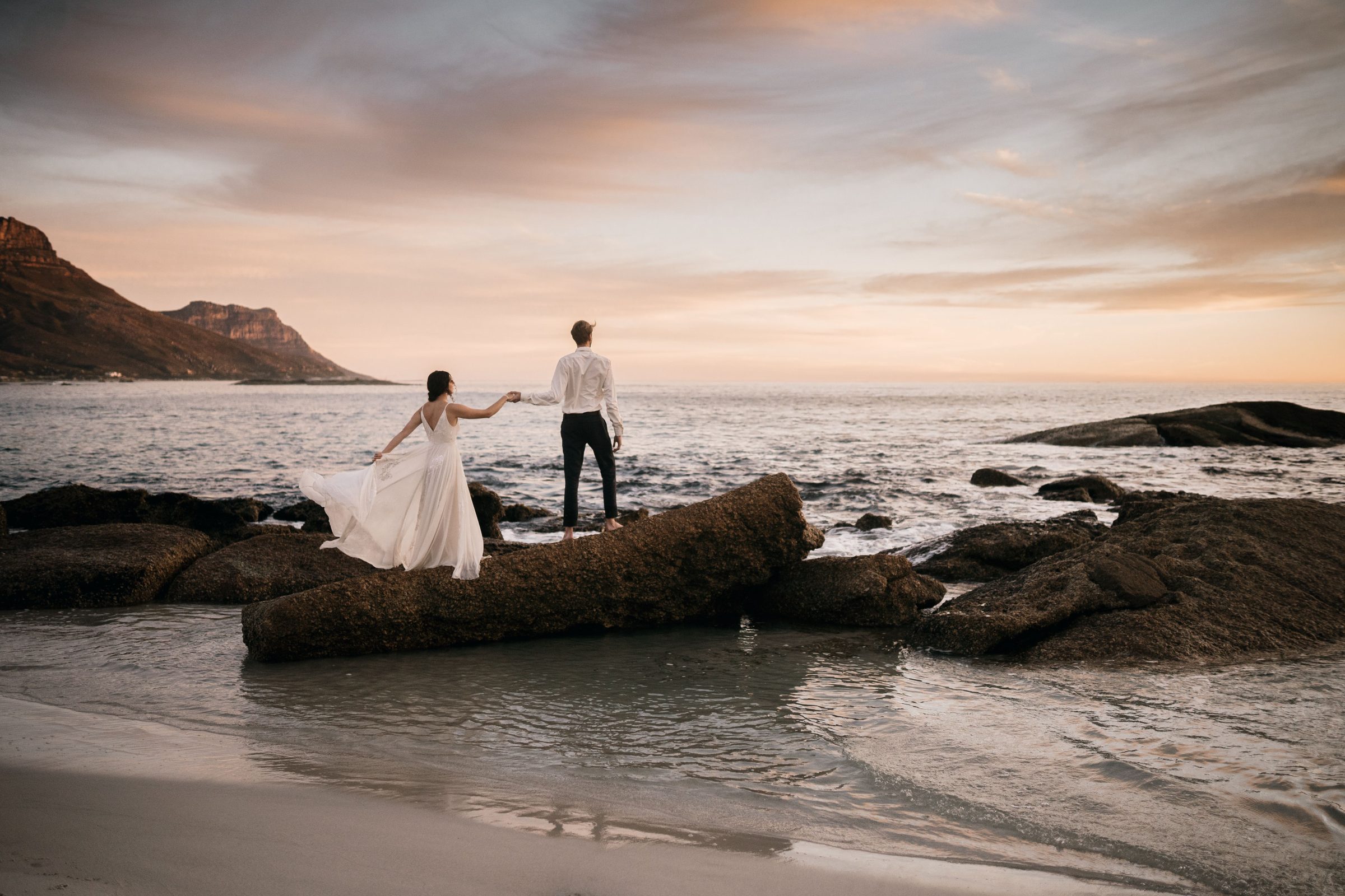 A bride and groom holding hands on the rocks at sunset on a Cape Town beach